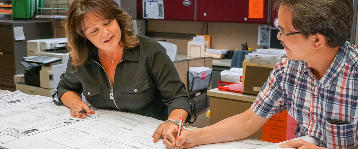 building counter picture of two people looking at a large map and discuss details. One is holding a pen while the other is looking at the map and pointing their finger.