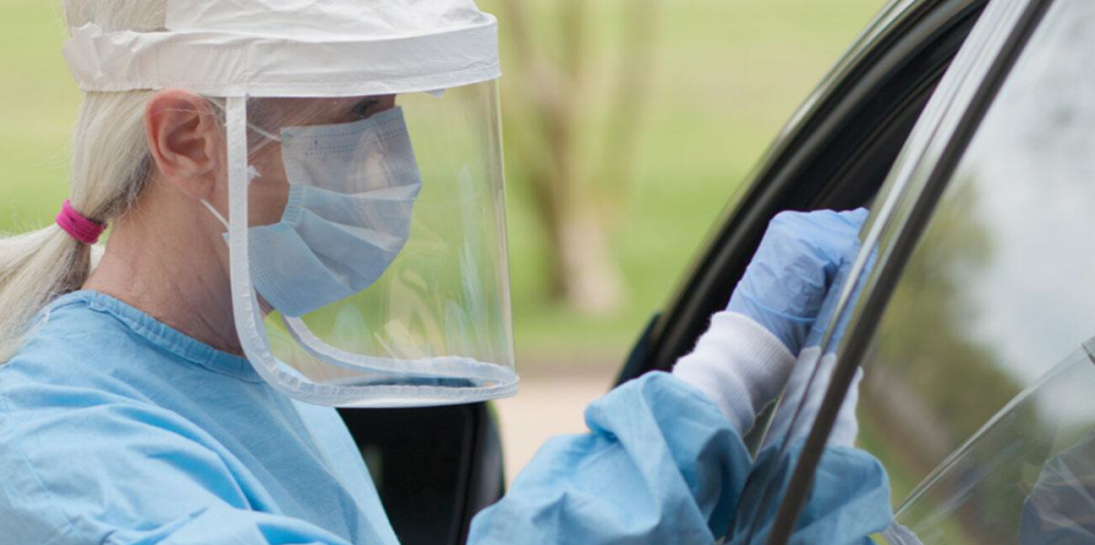 COVID-19 drive-through car testing site with a medical professional testing the driver while wearing a mask, a face shield, gloves, and a medical gown