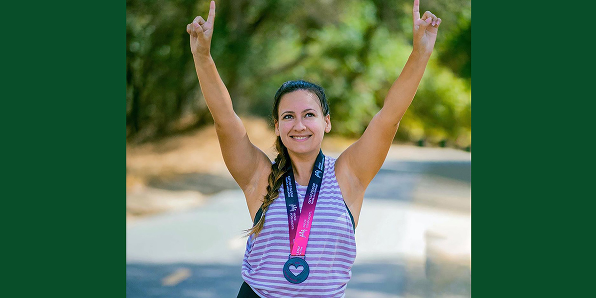 Love My Mom 2021 Photo of a racer wearing a colorful medal, a purple and white striped tank top, and braided hair, looking up with her hands pointed to the sky. 