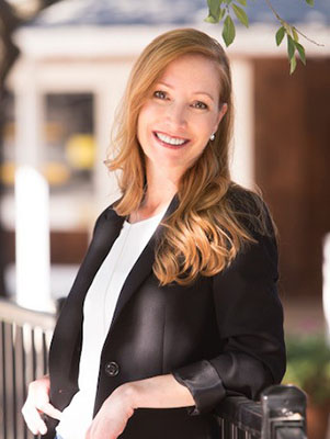 City Clerk Christa Freemantle Casual portrait headshot wearing a white shirt and a black unbuttoned blazer leaning on a black fence with a brown building with windows in the background