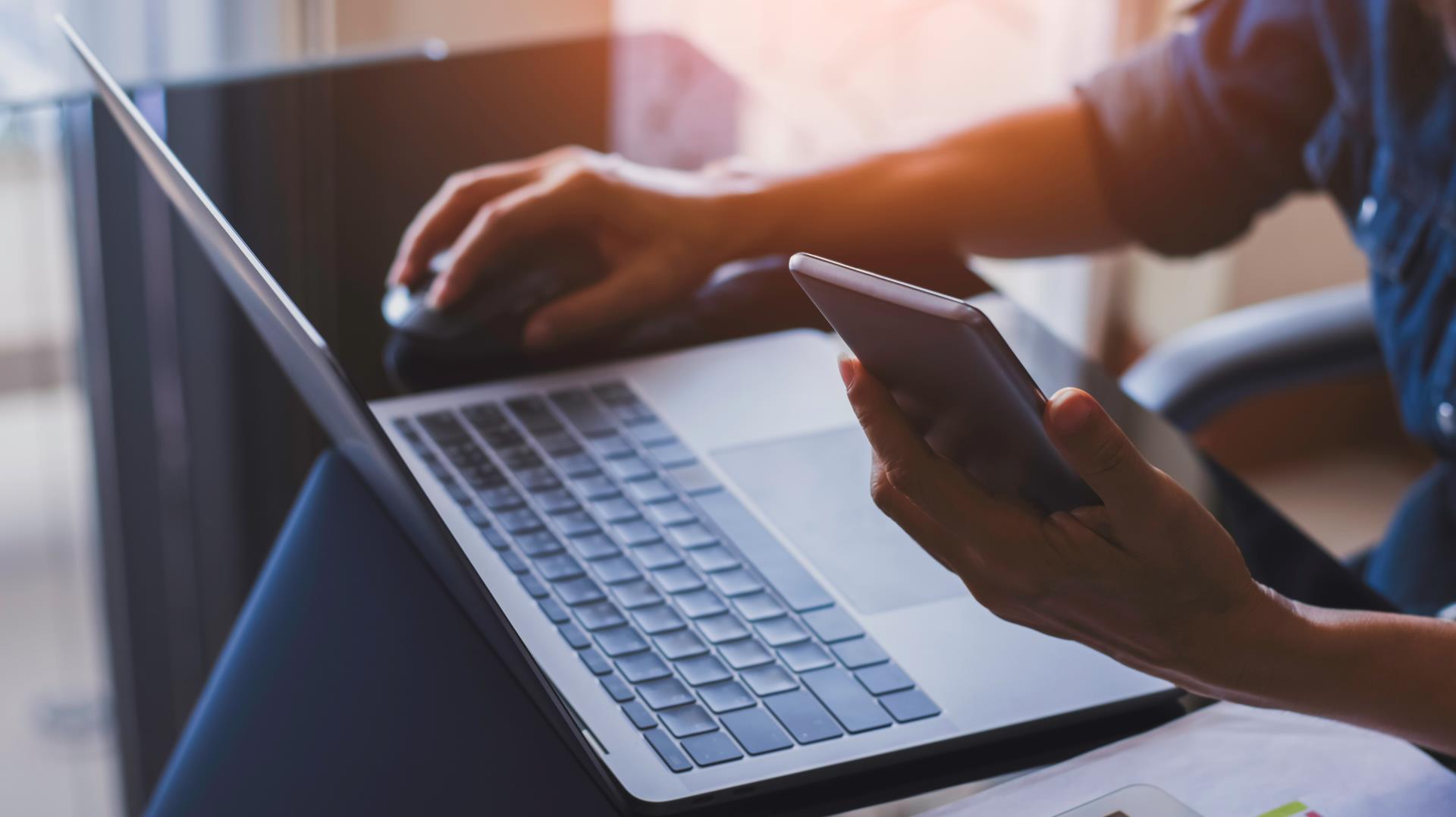 bill pay stock image of someone holding a mouse in one hand, a phone in the other hand, and a laptop in front of them with bills next to the laptop. Text says 