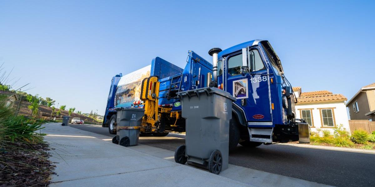 Refuse Truck - April 2021 photo from a low angle showing the truck using its arm to pick up a gray waste bin with other houses, broder park, and a blue sky in the background