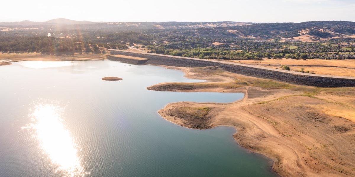 Folsom Lake July 2021 Drought-4 image of the lake reflecting the sun on the left and the exposed dirt area, trees, and neighborhoods on the right and in the background