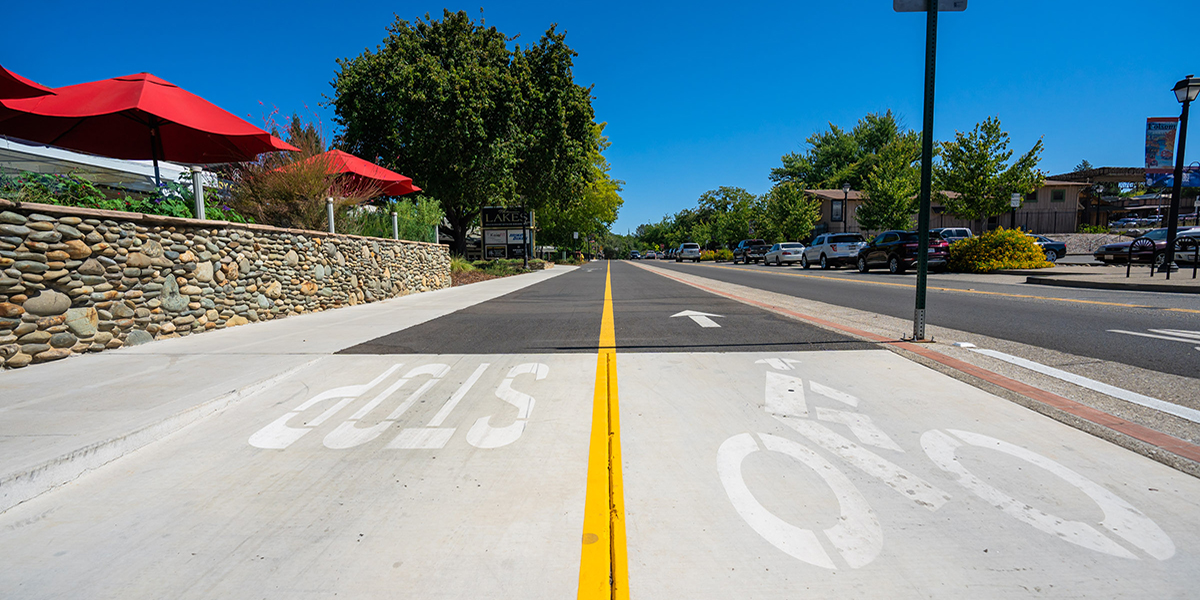 ATP virtual meeting Photo of Leidesdorf St. with a bright yellow divider for Stop and Bike lanes, with a patio dining with red umbrellas and green trees on the left and cars on the right