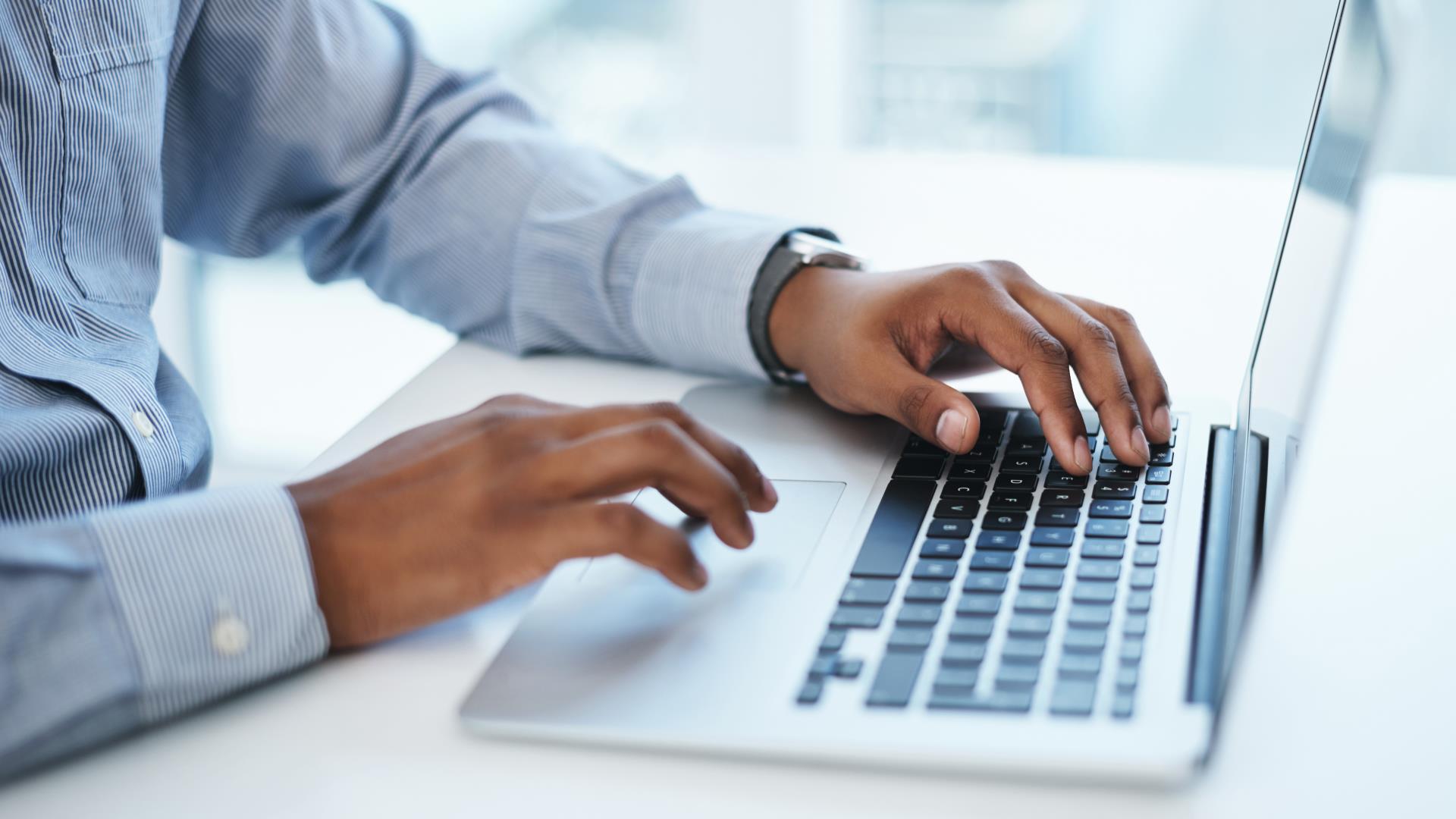 Getty Images of an employee wearing a blue and white pin striped button up long sleeve wearing a watch and working on a mac laptop