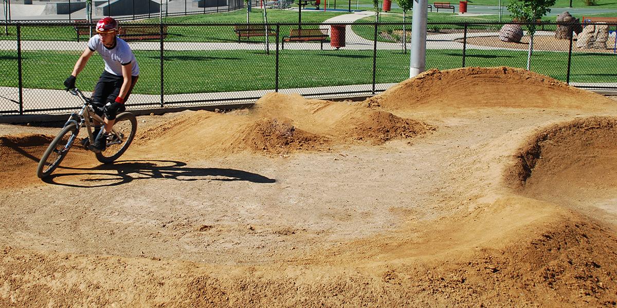 bike park dirt jump with hills and a cyclist on a bike wearing a red helmet, gray shirt, and black shorts turning, with park benches, grass, play structures, and the skate park in the background