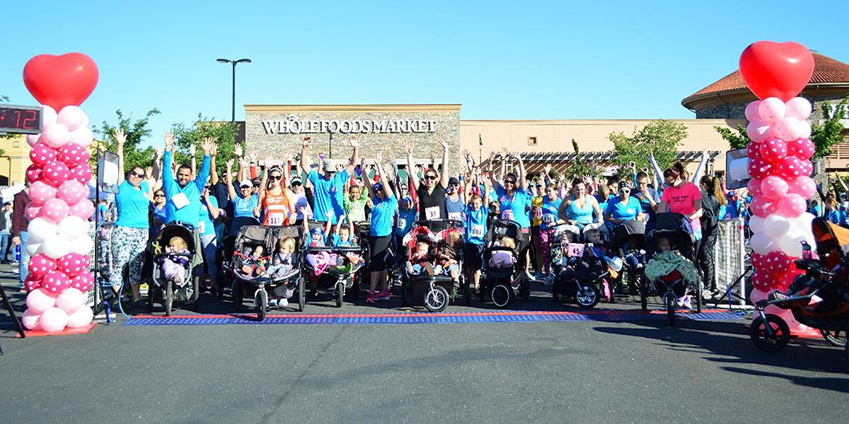 Love my mom race start with moms raising their hands with their children in strollers at the start of the line with two pink, red, and white balloon arches on either sides of the group