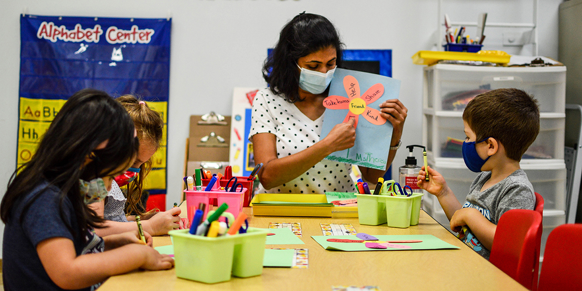 Preschool Open House 2022 with a teacher wearing a mask showing a handmade flower project with three masked students coloring their own flower projects