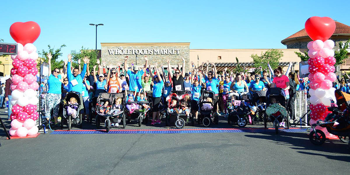 Love My Mom Start Line Cheer in front of Whole Foods Market with a clear blue sky and various pink balloon arches with participants raising their hands