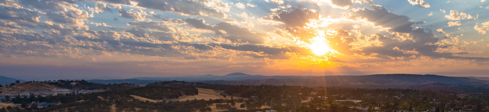 Early Folsom Sunrise banner overlooking the hills and trees with the orange bright sun flare in the corner and several clouds in the sky