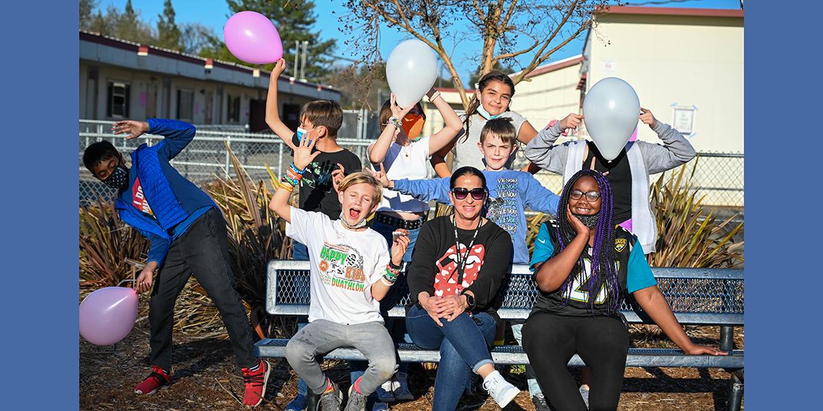 Summer CAVE kids and a teacher posing for a silly photo on the bench in front of their school with baloons