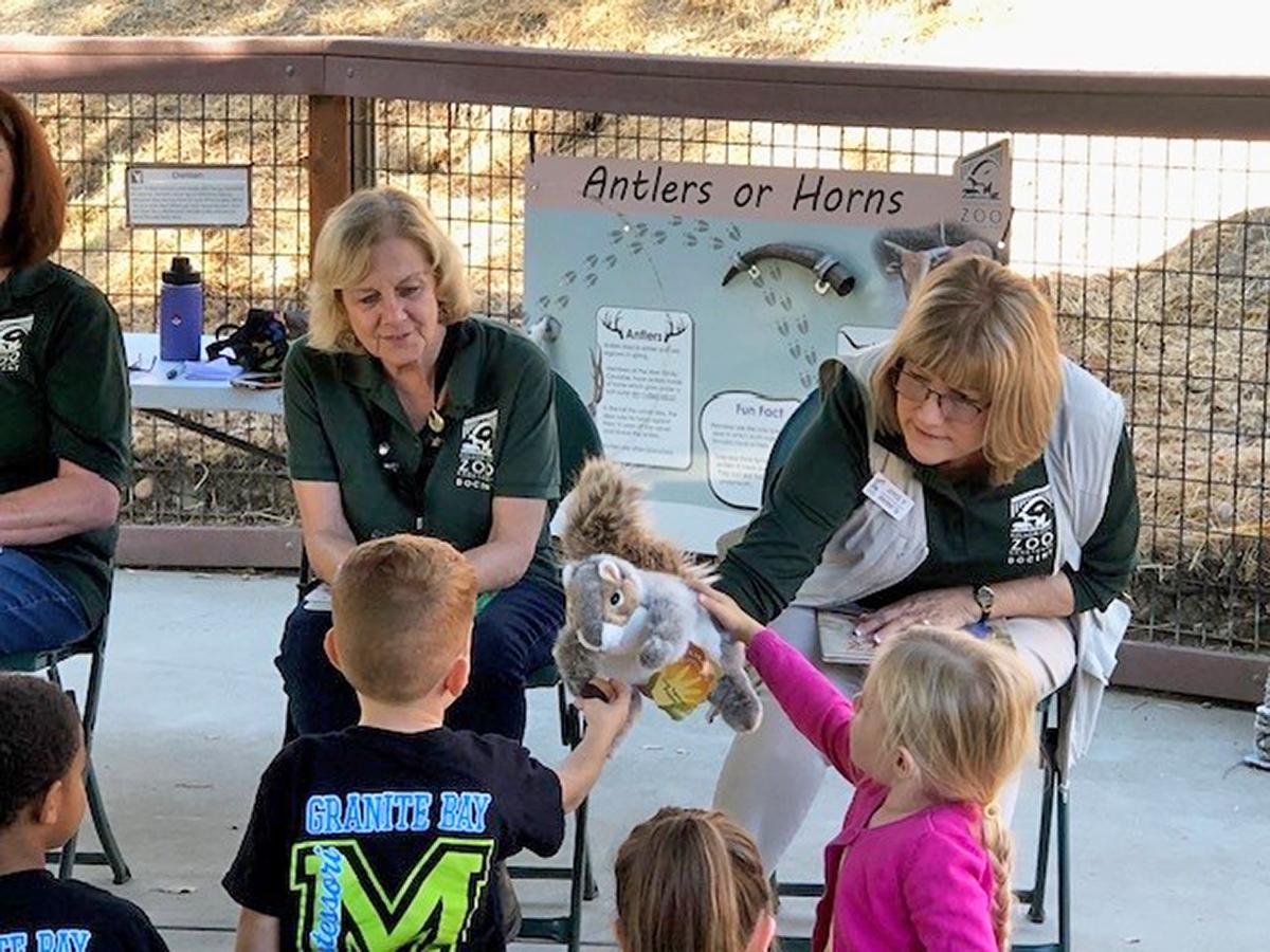 Two zoo docents sitting with children during a puppet storytime, one docent holding a squirrel puppet while the children listen attentively.