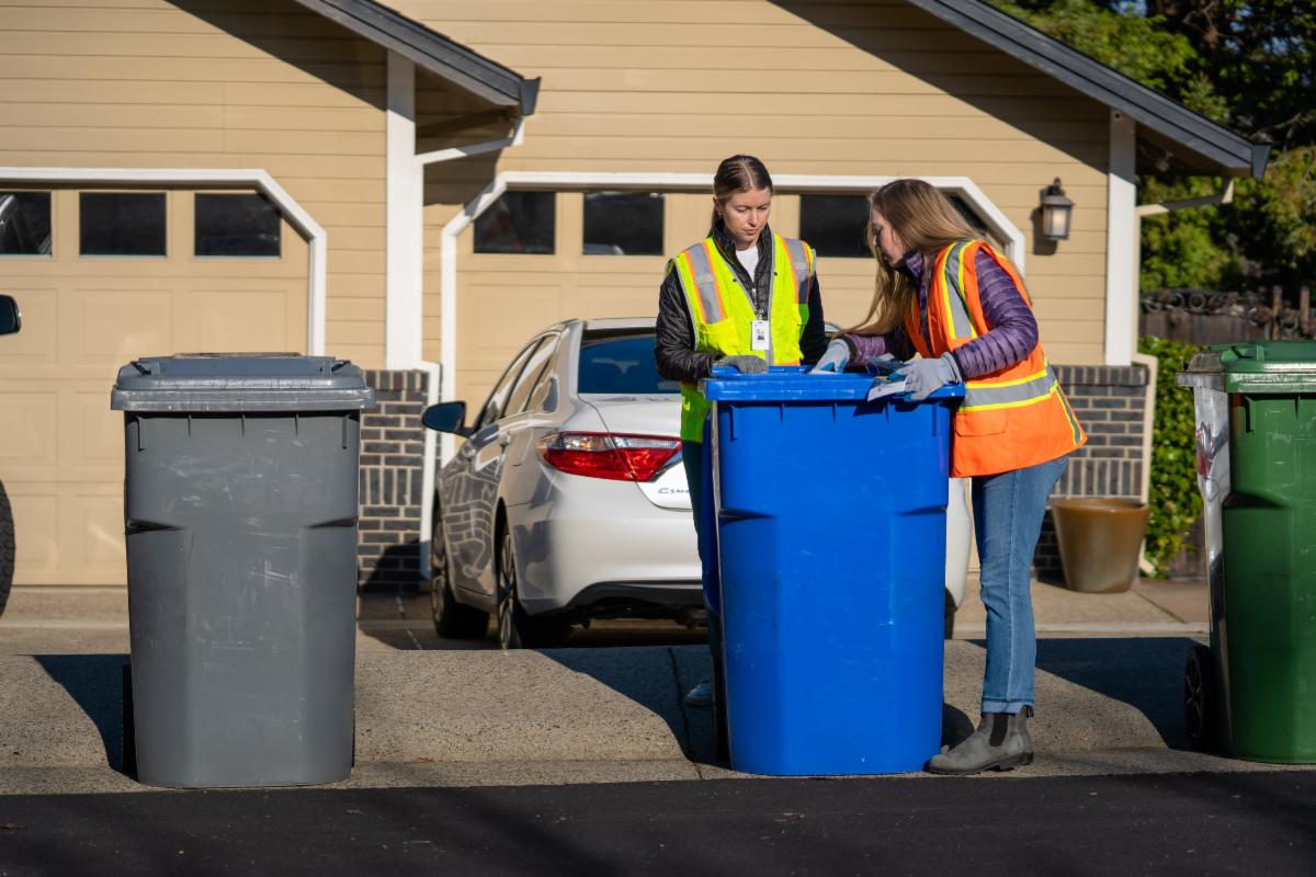 Two women in reflective safety vests inspecting a blue recycling bin on a residential street as part of a route review.