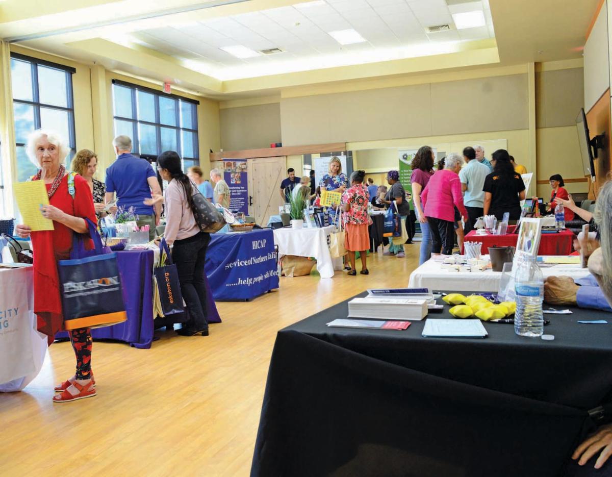 A busy community resource fair with seniors and other attendees visiting booths, interacting with representatives, and gathering information.