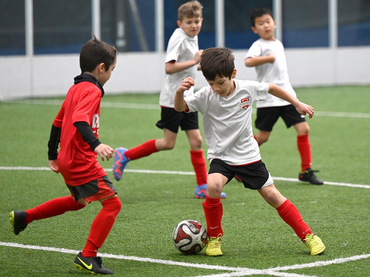 Boys in red and white uniforms playing soccer indoors, with one boy dribbling the ball while others attempt to intercept.