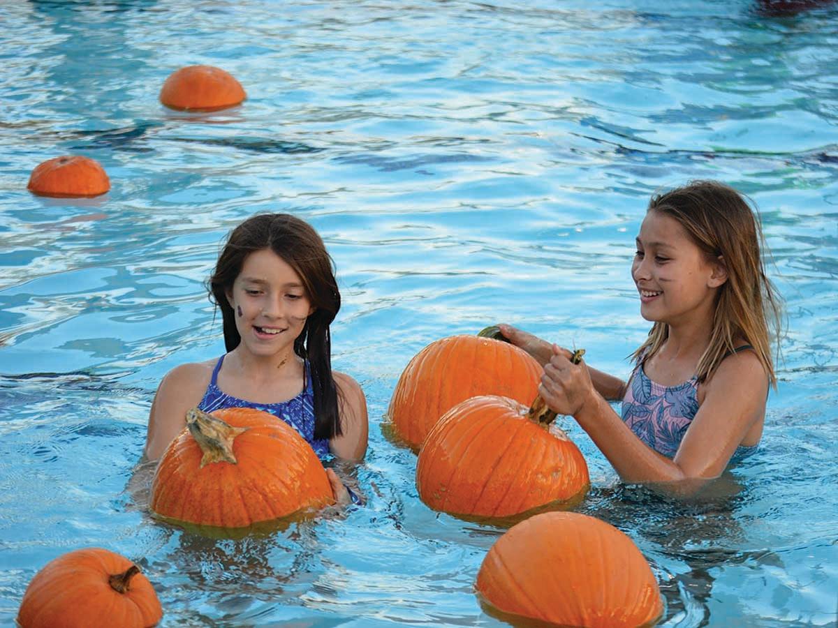 Two young girls swimming in a pool surrounded by floating pumpkins, smiling and enjoying the event.