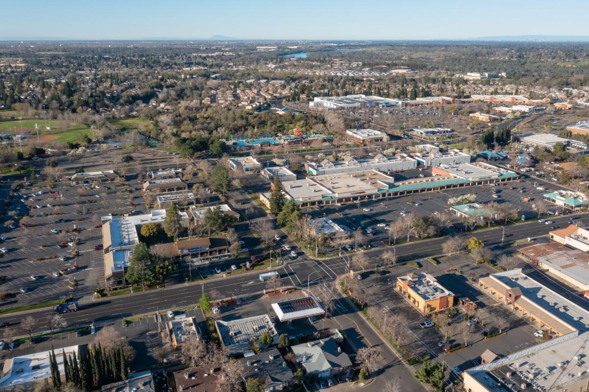 A drone shot of a business district, showcasing parking lots, buildings, and tree-lined streets under a clear sky.