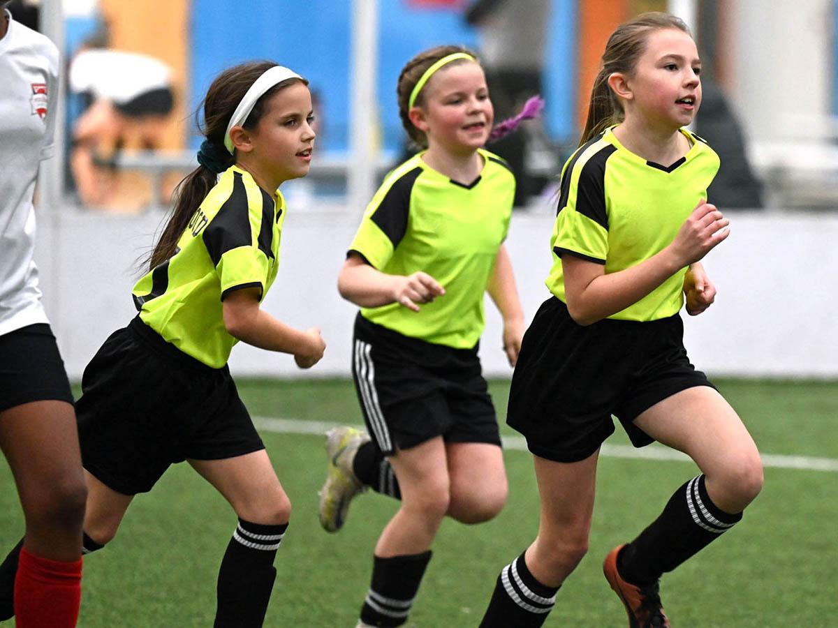 Three young girls wearing yellow and black uniforms are running during an indoor soccer game. They are focused on the game, with one girl leading, followed by two others close behind.