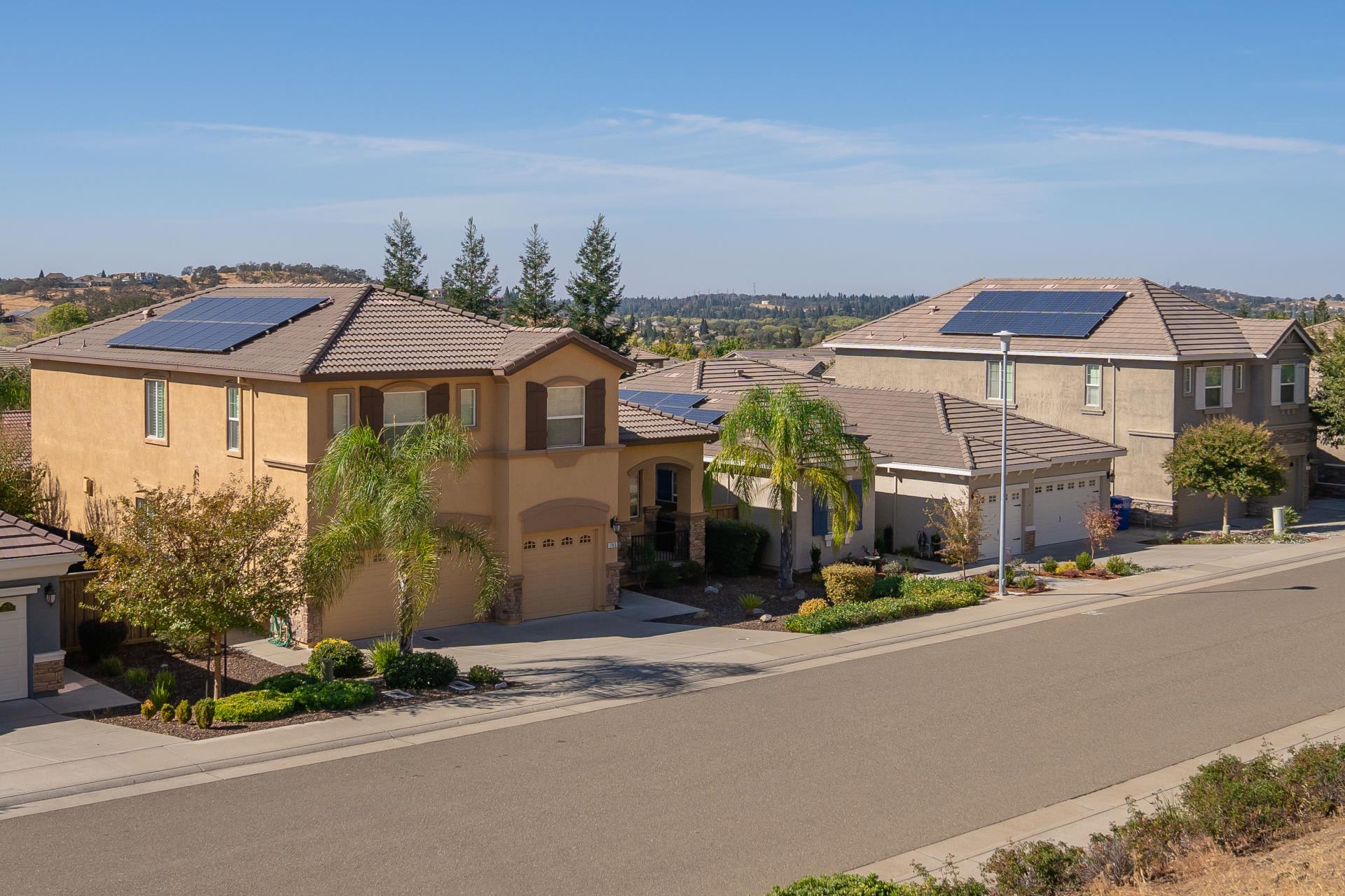 A row of modern two-story homes with solar panels installed on their roofs. The houses are surrounded by neatly landscaped yards, palm trees, and shrubs, with a clear blue sky and distant hills in the background.