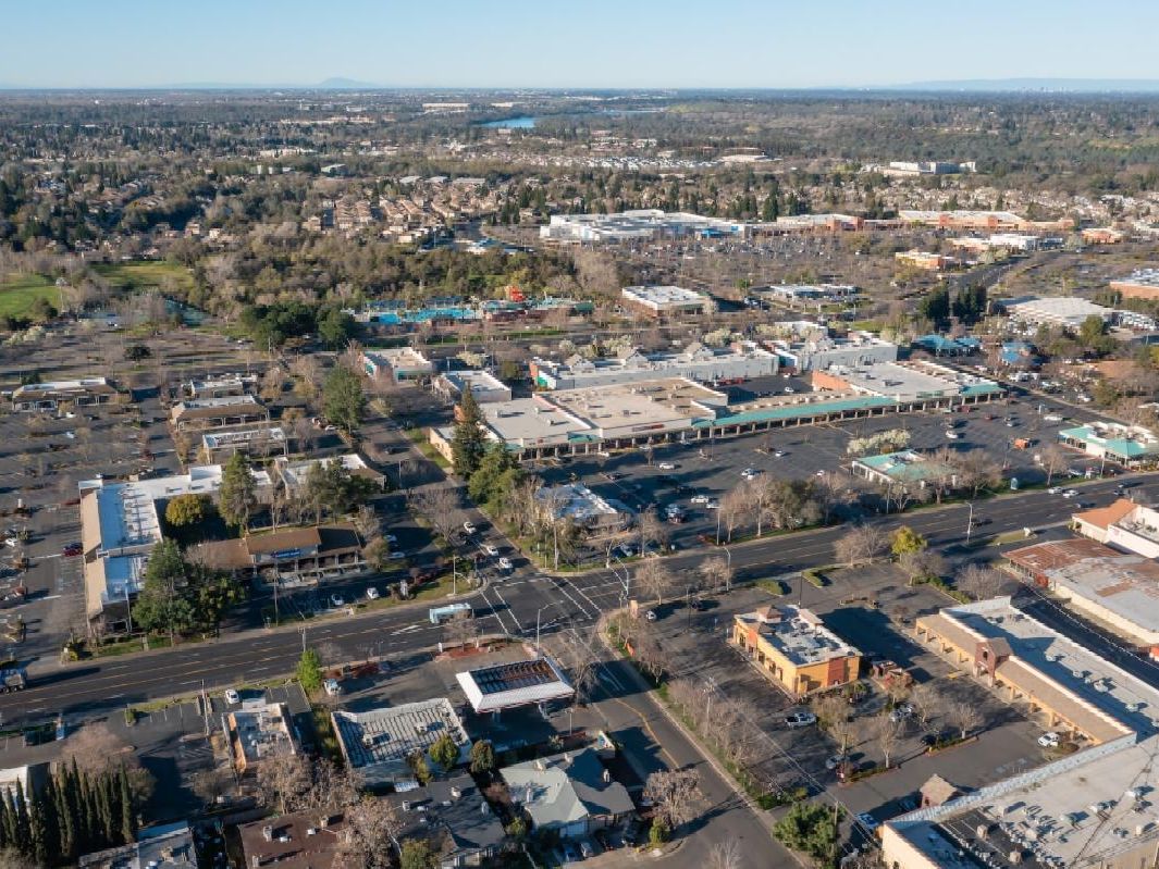 An overhead view of a city commercial district featuring parking lots, various buildings, tree-lined streets, and residential areas in the background.