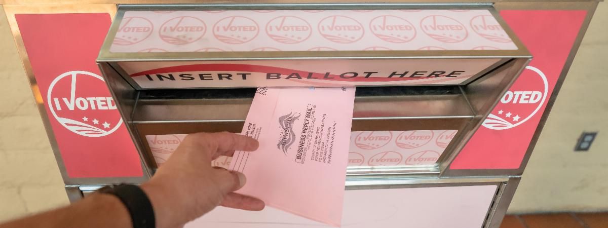 A close-up of a hand inserting a pink mail-in ballot into a labeled 