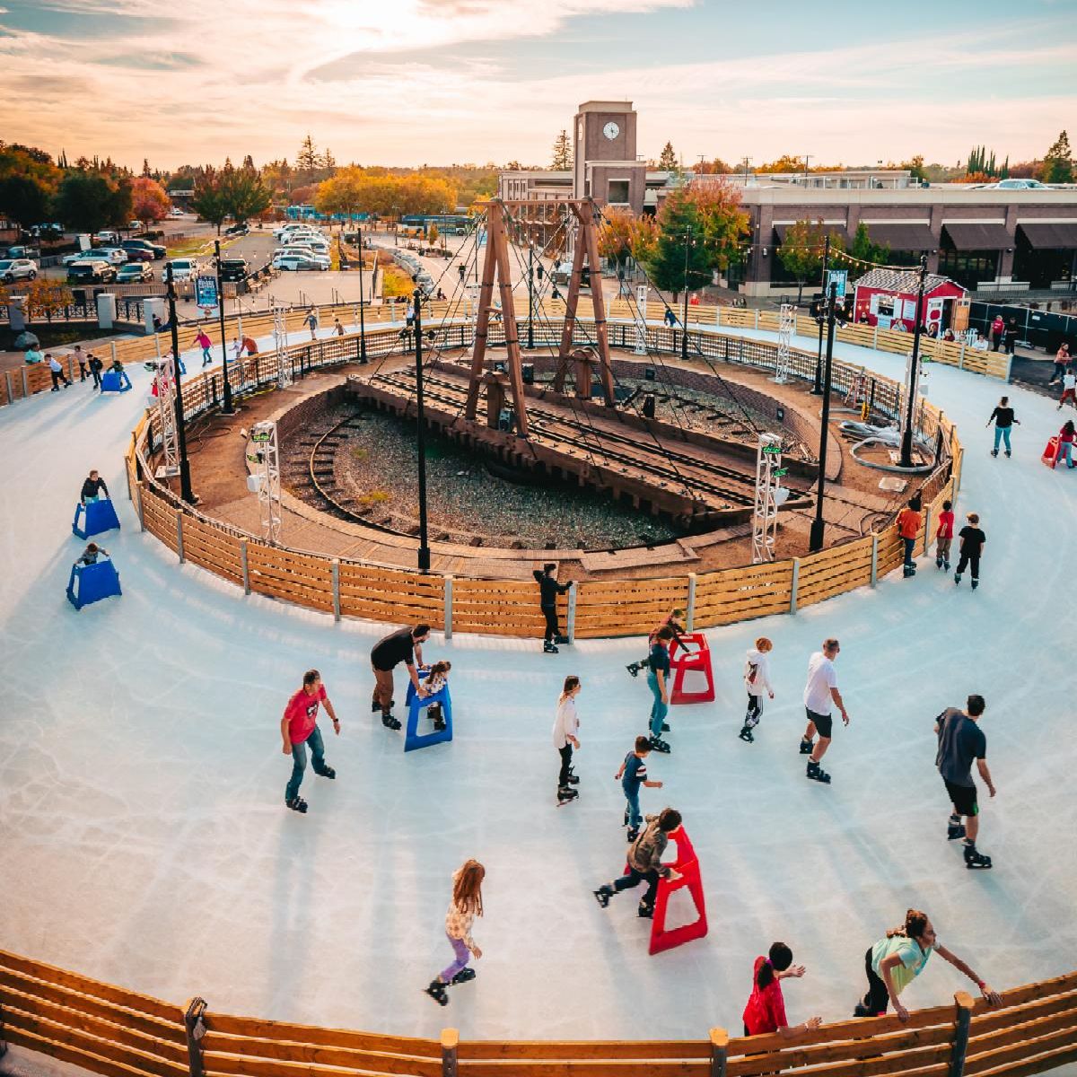 An aerial view of people skating around a circular outdoor ice rink with a central wooden structure, surrounded by a low wooden fence, trees, and a sunset-lit parking area.