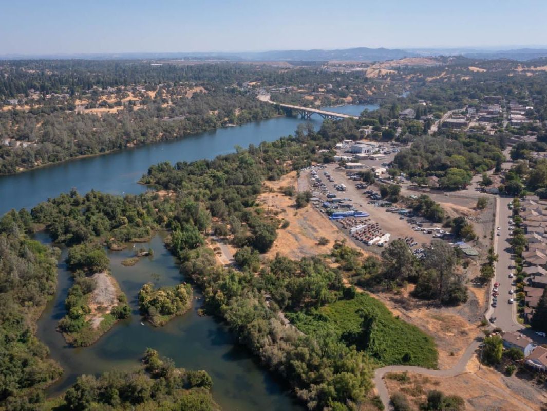 A scenic aerial view showing a river winding through a green landscape with nearby buildings, parking areas, and roads stretching toward distant hills under a clear sky.